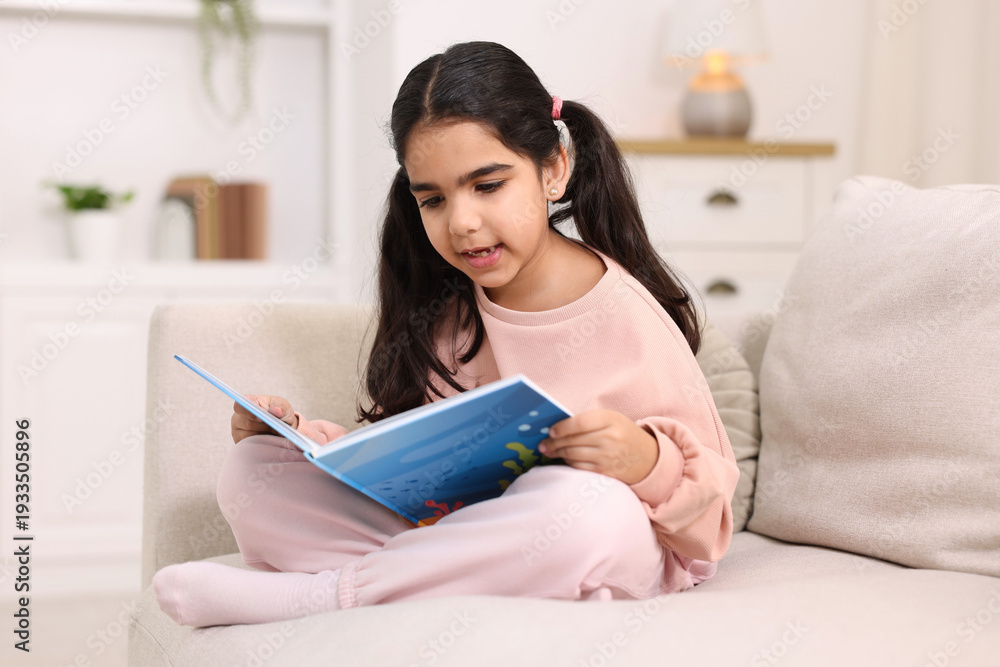 Naklejka premium Cute little girl reading book on couch at home