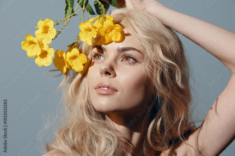 Naklejka premium Young woman with long blonde hair holding yellow flowers, embodying natural beauty and vibrant emotions against a blue background.