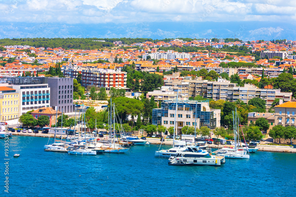 Obraz premium Harbor of Zadar, Croatia, numerous sailboats and yachts are moored in blue waters. Background features a sprawling cityscape of terracotta roofs and modern buildings set against distant mountains