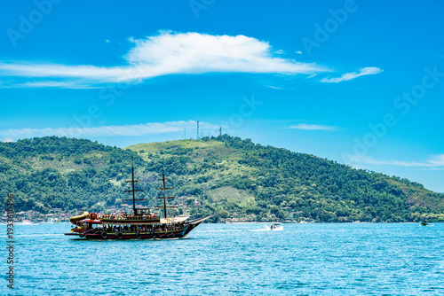 ketch-rigged wooden tourist boat sailing along Angra dos Reis coastline, Brazil. Features rainforest-covered mountains and traditional sailing charm for travel and documentary projects.