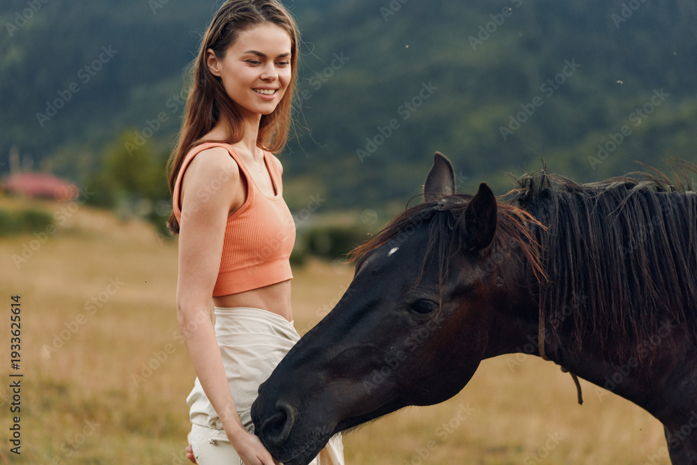 Obraz premium A smiling young woman stands in a sunlit field beside a dark horse, wearing an orange crop top and light shorts. The outdoor moment radiates calm, friendship, and natural beauty.
