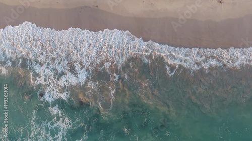 Aerial top down view of ocean wave breaking along sandy tropical shore with smooth left to right movement