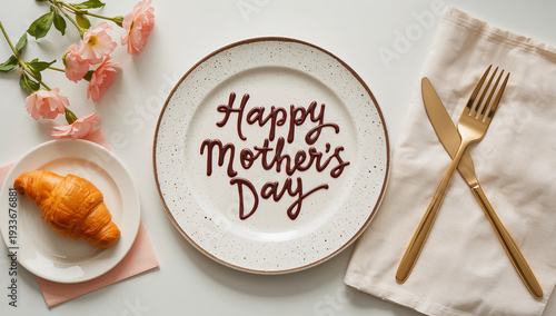 A single croissant on a plate next to a formal setting with 'Happy Mother's Day' written in chocolate on a larger plate.