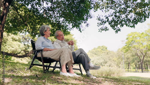 A happy senior Asian couple sits on folding chairs under a shady tree in a lush park. They enjoy a peaceful conversation, showcasing a healthy retirement, lasting love, and wellness in nature