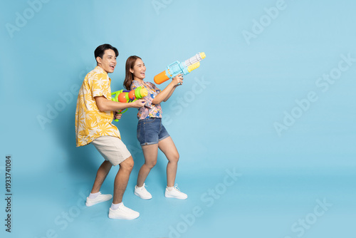 A cheerful Asian couple in floral shirts aims colorful water guns, celebrating the Songkran Festival with laughter against a blue background, Thai New Year and summer travel themes