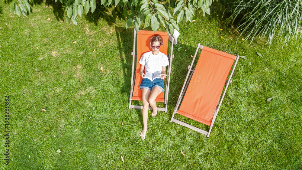Fototapeta premium Young girl relaxes and reading book in summer garden in sunbed deckchair on grass, relaxing outdoors in green park on weekend, aerial drone view from above