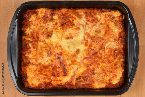 A top view of a freshly baked lasagna with a golden brown cheese crust and rich meat sauce served in a black baking tray on a wooden surface