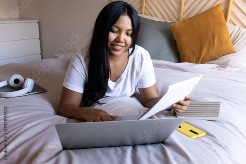 Fotografie Young latina woman working from home on bed using laptop and reading documents