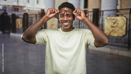 Photos Man adjusts eyeglasses with both hands at temples, smiling broadly in front of ornate railings inside a public building; joyful confidence