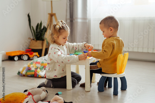 Two children work together at a table, building with colorful blocks. The room is bright and has toys scattered on the floor
