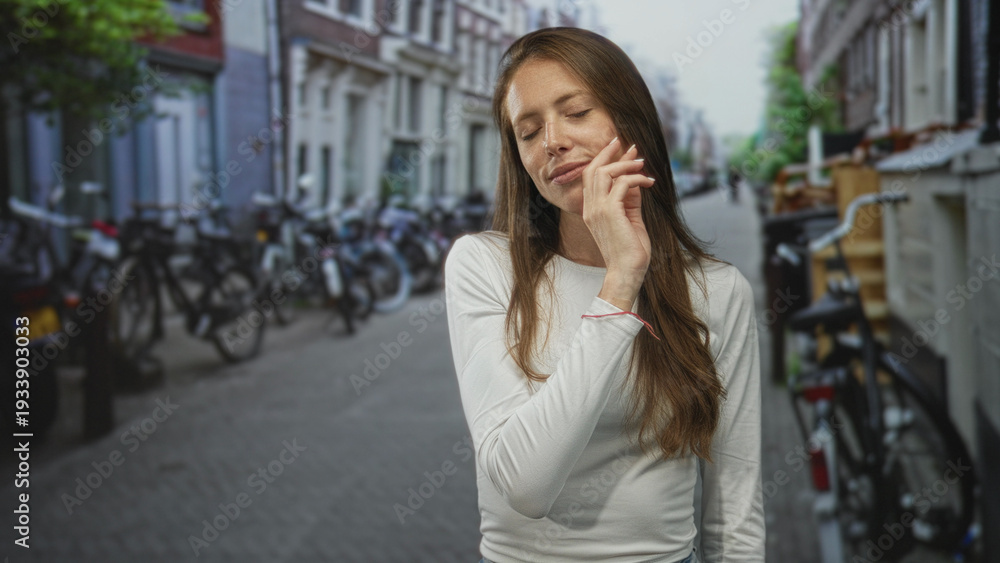 Fototapeta premium Young hispanic woman touching cheek with right hand while smiling gently on a narrow city street lined with bicycles; serenity.