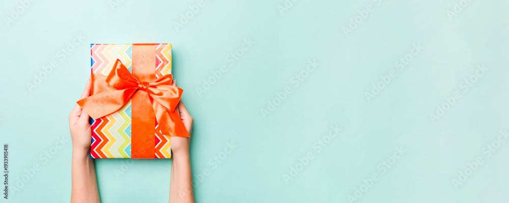 Fototapeta premium Woman hands give wrapped christmas or other holiday handmade present in colored paper. Present box, decoration of gift on Blue table, top view with copy space