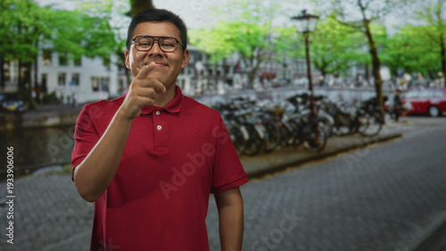 Man in red polo adjusting glasses on cobblestone street by a canal with parked bicycles and lamppost; youthful confidence.