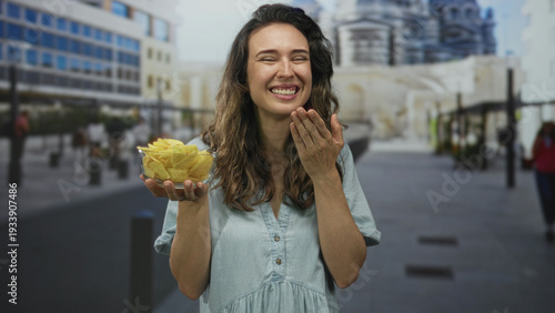 Woman smiling and holding glass bowl of crispy chips with hand to mouth gesture on street; happiness.