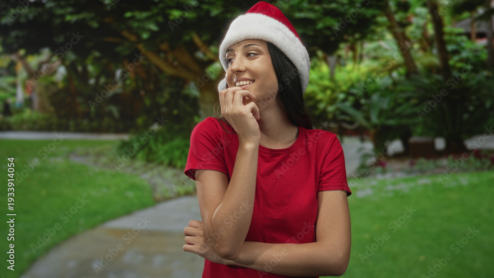 Fototapeta premium Woman biting nails with hand to mouth on park path street wearing santa hat and red shirt; playful thoughtfulness.