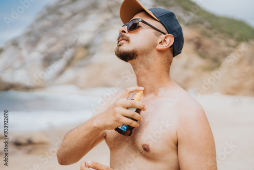 A man wearing a cap and sunglasses stands on a sunlit beach, spraying sunscreen. He looks relaxed, enjoying the seaside scenery with rocky cliffs in the background, capturing coastal summer vibes.