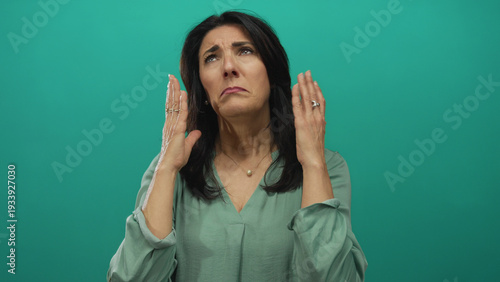Valokuva Middle aged woman raises hands to face in teal studio setting with expressive gesture and closed eyes showing despair; frustration