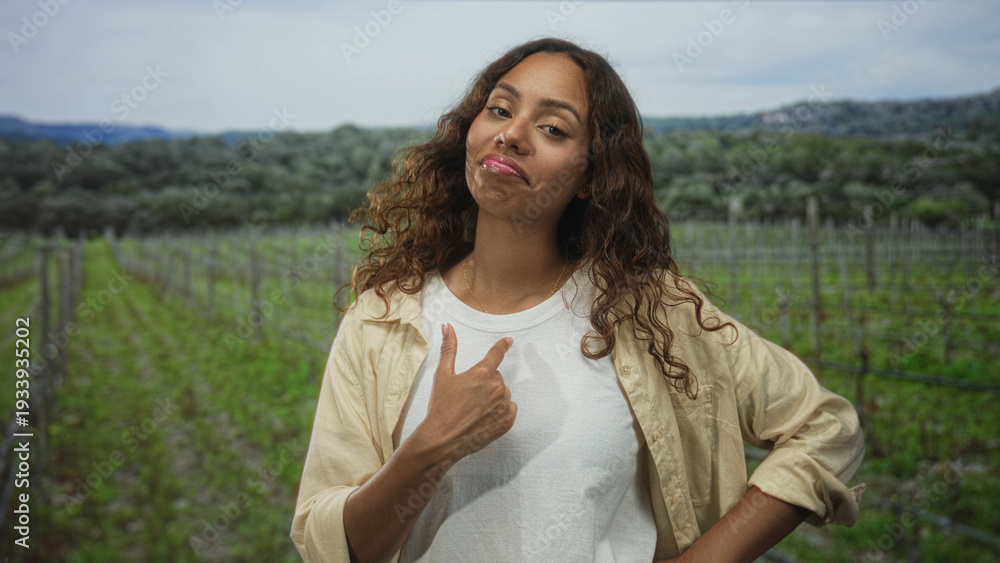 Naklejka premium Woman points thumb to chest in a vineyard field, smiling with hand on hip and wearing white t shirt and beige overshirt; confidence.