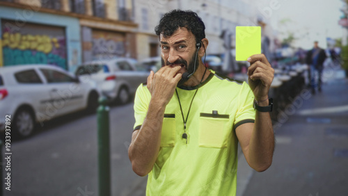 Fotografie Man referee middle age hispanic with beard bites nails with hand while holding yellow card and whistle headset on busy city street; anxiety hesitation