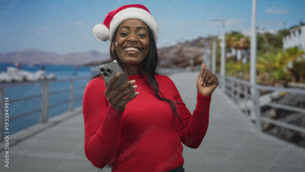 Fototapeta premium Woman wearing santa hat and red sweater holds smartphone, smiles and raises fist on street boardwalk by water; holiday joy celebration.