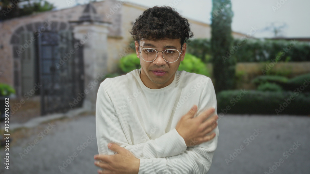 Fototapeta premium Young man hugs bare shoulders outside stone building entrance as he shivers in light sweater; anxiety.