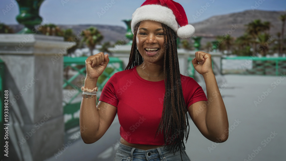 Fototapeta premium Woman in red shirt and santa hat smiles and raises clenched fists on street with green railing and palm trees by the sea; holiday joy.