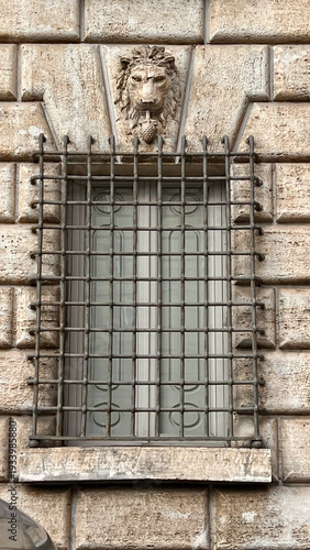 Architectural detail of historic stone window with iron grid and lion sculpture decoration on building facade in Rome, Italy.