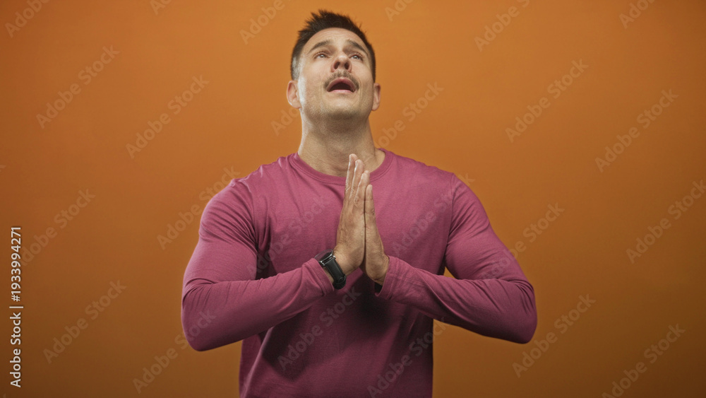 Obraz premium Hispanic man with mustache presses hands together in prayer, head tilted up in a studio with orange backdrop; hope faith gratitude devotion.