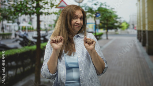 Φωτογραφία Woman with hands clenched near chest in a recoil gesture on a city street with sidewalk and parked scooters; unease caution