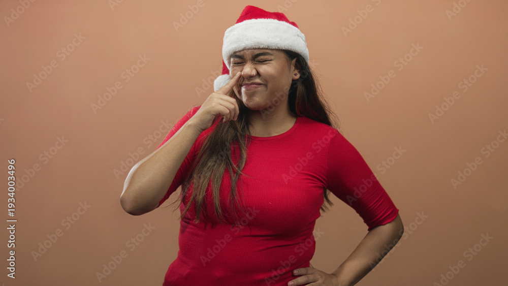 Fototapeta premium Woman pointing finger to nose wearing santa hat in studio, red shirt and hand on hip with subtle smile and nose piercing; playful holiday cheer.
