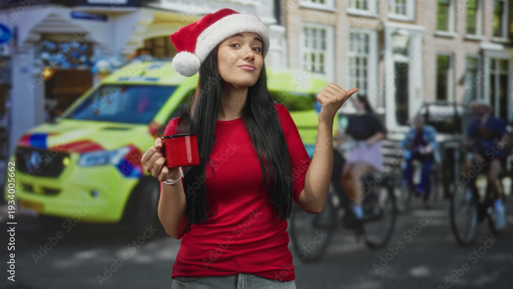Fototapeta premium Young woman in santa hat holding red mug, thumb pointing to street with emergency vehicle behind; holiday cheer.
