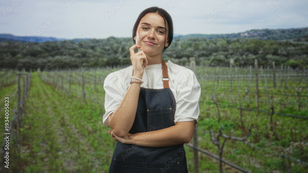 Fototapeta premium Woman in apron touches chin with hand, arms folded, standing among vineyard rows in forest setting with distant hills and olive trees; calm thinking.