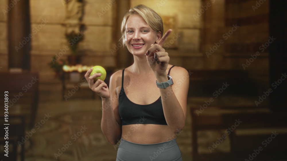Fototapeta premium Woman holding green apple, finger to lips for silence in building wearing sports bra and leggings; playful tease.