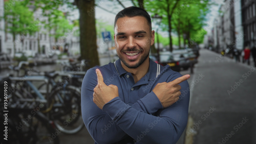 Fototapeta premium Smiling hispanic man pointing index fingers across his chest on city street near parked bicycles and cars; choice confidence.