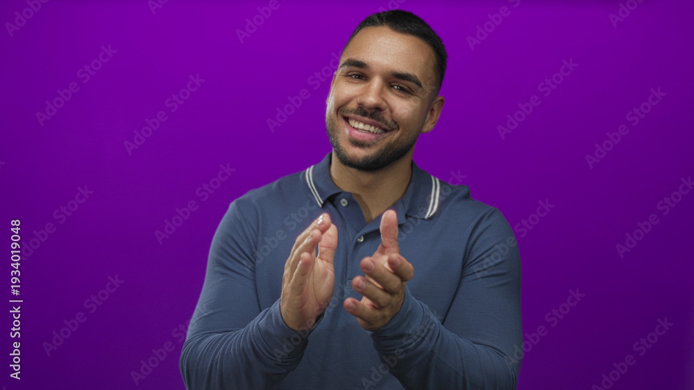 Fototapeta premium Young hispanic man wearing a blue polo shirt claps hands with a broad smile in a purple studio setting; celebration.