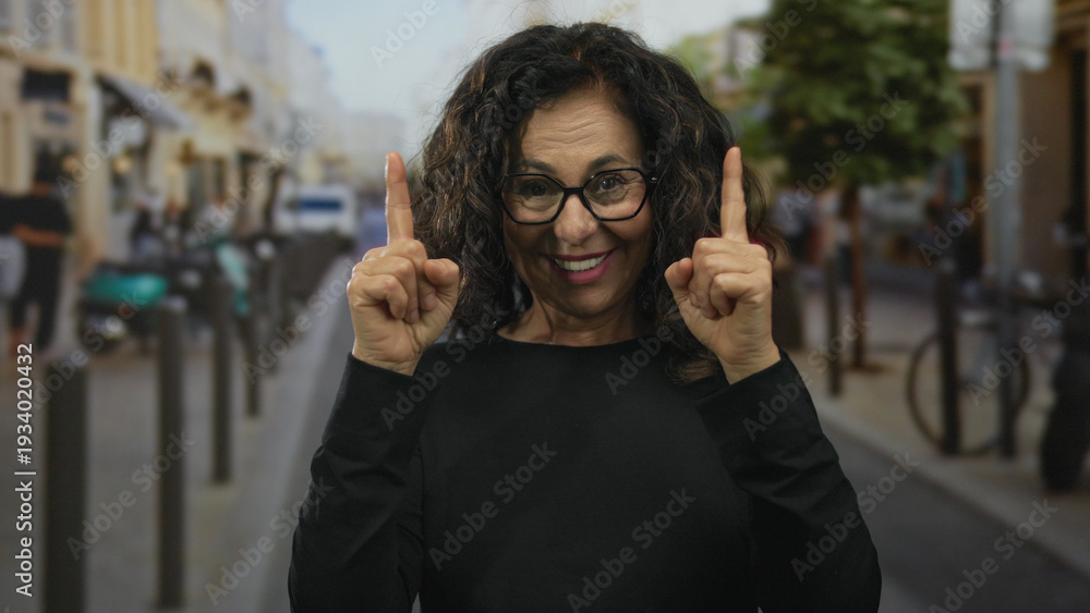 Fototapeta premium Middle aged hispanic woman points both index fingers upward on street while smiling amid bustling storefronts; hope optimism aspiration joy.