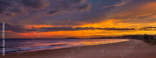 Wide panoramic view of Narrabeen Beach at sunset with surfers on sand. Dramatic orange sky over Northern Beaches coastline in Sydney Australia. Perfect for travel and vacation marketing.