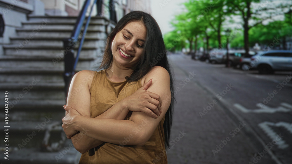 Fototapeta premium Woman hugs herself with bare arms on a city street by stone stairs, smiling in a casual pose and closing eyes; self care joy.