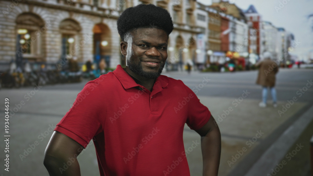 Obraz premium Man with hands on hips wearing red polo on street near building, smiling and posing; urban confidence.