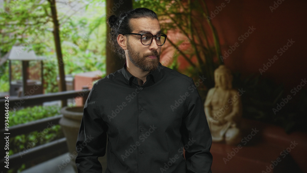 Obraz premium Man with glasses and beard standing in a hotel building lobby, wearing black shirt and gazing sideways near balcony and statue; contemplation.