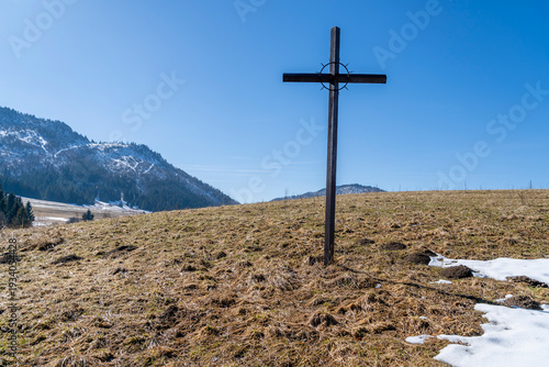 A simple steel cross with a crown of thorns in spring in a clearing with mountains in the background.