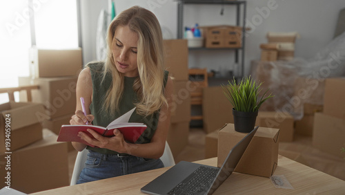 Canvastavla Woman writing in notebook in living room surrounded by moving boxes and laptop, indicating new home environment and planning process