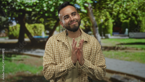 Man smiling with hands pressed together and tilted head in forest path among green trees, casual checked shirt visible; calm gratitude.