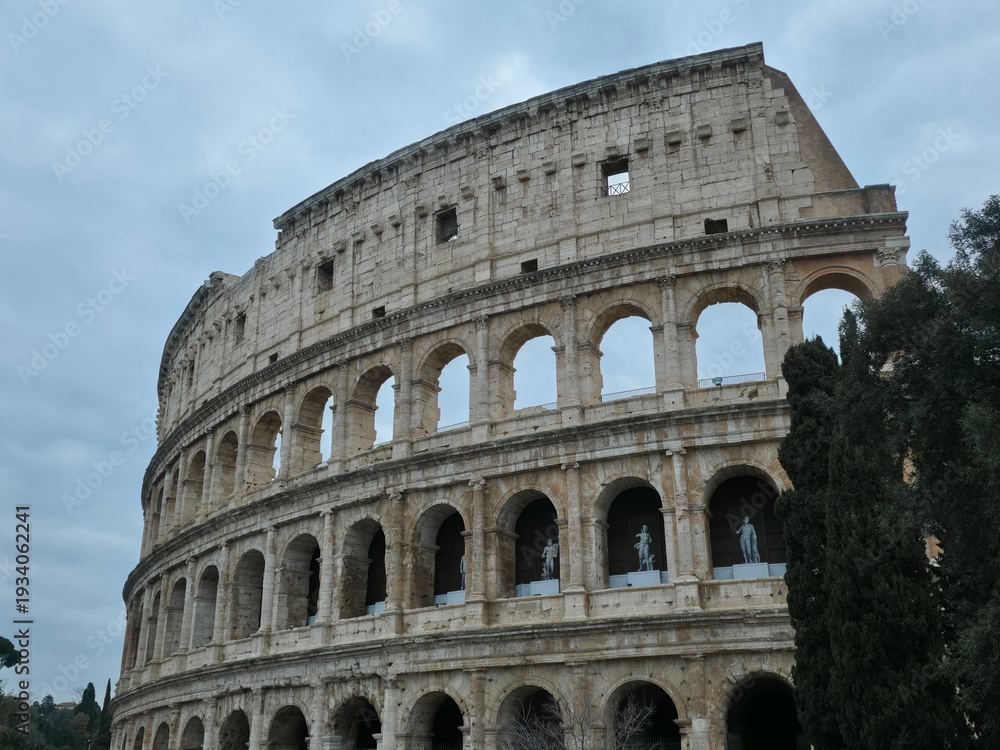Obraz premium Ancient Roman Colosseum exterior with stone arches and classical statues, iconic Flavian Amphitheatre landmark under overcast sky, Rome, Italy