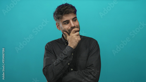 Photos Young hispanic man wearing dark shirt with hand on chin and intense gaze against turquoise wall in studio; reflective