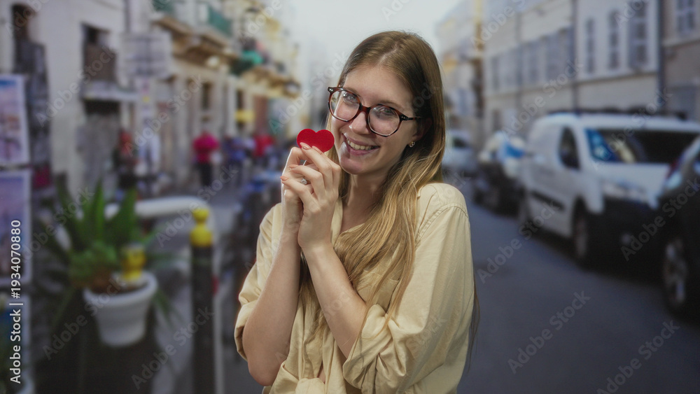 Obraz premium Young blonde woman wearing glasses and smiling while holding a small red heart on a busy street; love.
