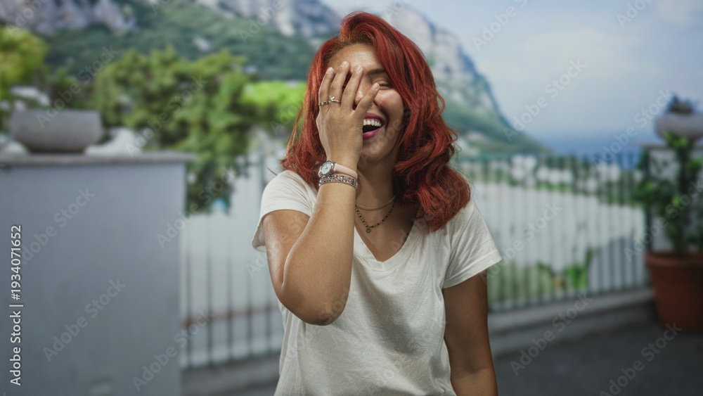 Fototapeta premium Woman laughing with hand covering face on building balcony wearing white t shirt, red hair and watch, rings visible; joyful.