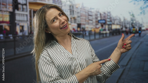 Free Download Woman with long blonde hair pointing finger down sunlit busy city street with blurred storefronts and tram rails in view; joy. - GFXLoad