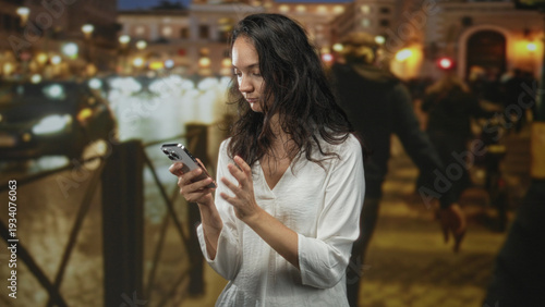 Photos Woman holding smartphone with arms crossed on a busy night street by waterfront railing, gazing to the side; solitude contemplation