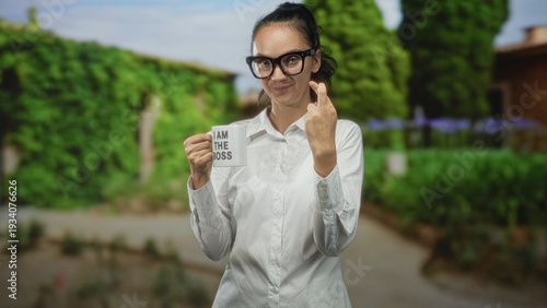 Woman wearing glasses and white shirt holding mug reading i am the boss, raises middle finger while standing by building; defiance.